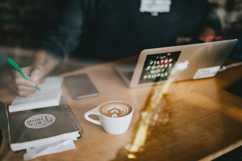 Developer belajar coding secara mandiri di kafe dengan laptop dan catatan — belajar sendiri vs bootcamp