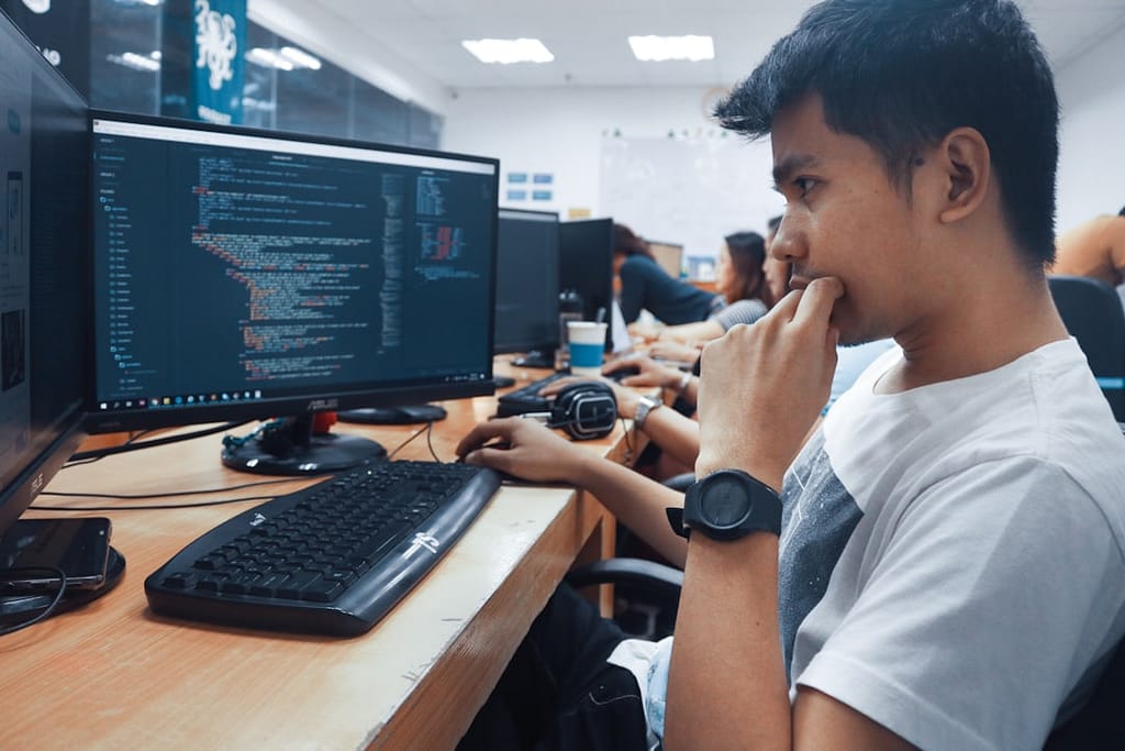 Seorang mahasiswa muda sedang belajar coding dengan melihat layar komputer berisi kode, suasana kelas atau lab komputer