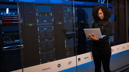 Cloud engineer perempuan bekerja dengan laptop di depan server rack di data center modern. Photo by Pexels.